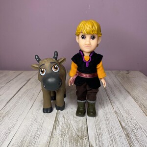 K&ouml;nnte beinhalten: Eine Puppe einer Figur mit blonden Haaren, die eine schwarze Weste &uuml;ber einem orangefarbenen Hemd, braune Hosen und gr&uuml;ne Stiefel tr&auml;gt. Eine braune Rentierfigur steht neben der Puppe. Die Figuren stehen auf einer wei&szlig;en Holzoberfl&auml;che.