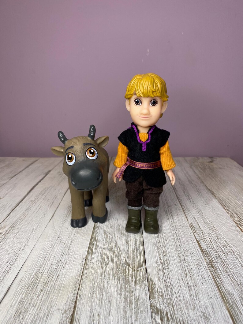 K&ouml;nnte beinhalten: Eine Puppe einer Figur mit blonden Haaren, die eine schwarze Weste &uuml;ber einem orangefarbenen Hemd, braune Hosen und gr&uuml;ne Stiefel tr&auml;gt. Eine braune Rentierfigur steht neben der Puppe. Die Figuren stehen auf einer wei&szlig;en Holzoberfl&auml;che.