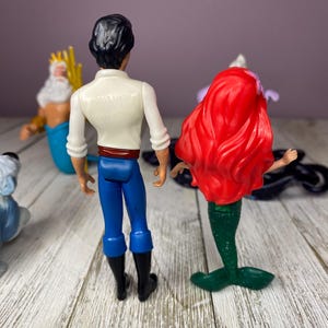 May include: A collection of Disney figurines featuring characters from The Little Mermaid. The figures include Prince Eric in blue pants and black boots, Ariel with red hair and a green tail, and King Triton with a golden trident.