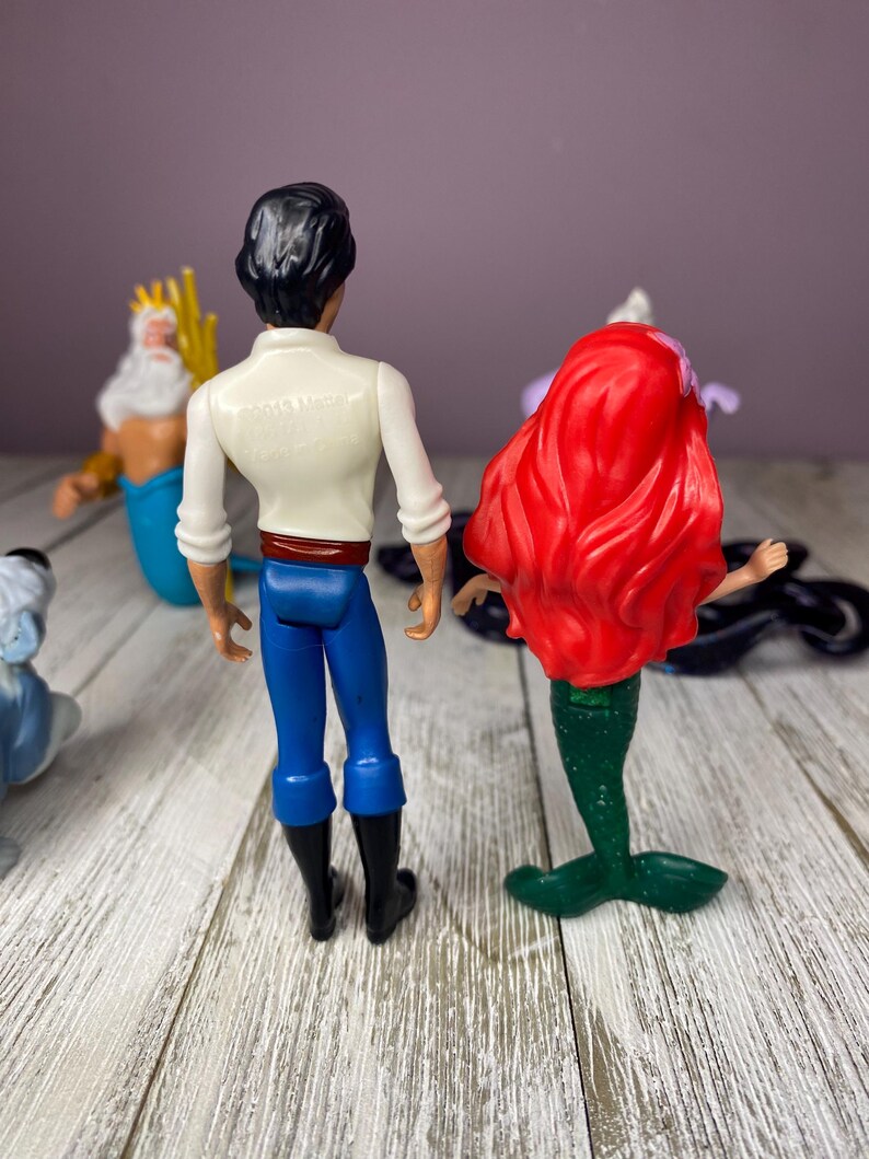 May include: A collection of Disney figurines featuring characters from The Little Mermaid. The figures include Prince Eric in blue pants and black boots, Ariel with red hair and a green tail, and King Triton with a golden trident.