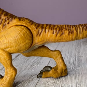May include: Close-up of a plastic dinosaur toy in shades of yellow and brown. The toy features detailed textures and markings, with a focus on the leg and tail. The text "MADE IN VIETNAM" is visible on the toy.