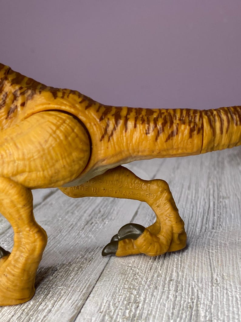 May include: Close-up of a plastic dinosaur toy in shades of yellow and brown. The toy features detailed textures and markings, with a focus on the leg and tail. The text "MADE IN VIETNAM" is visible on the toy.