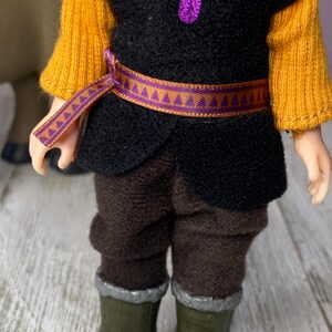 K&ouml;nnte beinhalten: Eine Puppe in einem Kost&uuml;m mit schwarzer Weste, senfgelben &Auml;rmeln und brauner Hose. Die Puppe hat einen lila G&uuml;rtel und eine passende lila Halskette. Die Puppe tr&auml;gt gr&uuml;ne Stiefel mit silberner Verzierung.