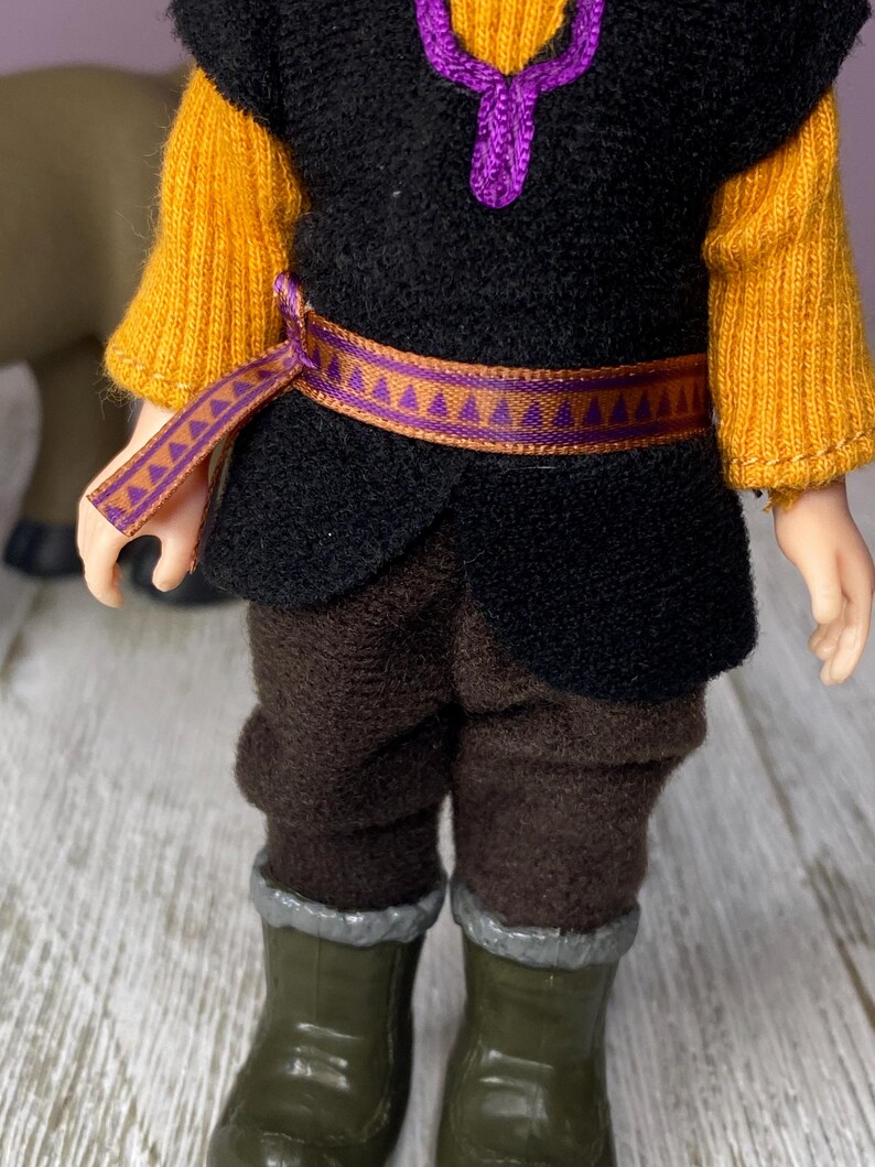 K&ouml;nnte beinhalten: Eine Puppe in einem Kost&uuml;m mit schwarzer Weste, senfgelben &Auml;rmeln und brauner Hose. Die Puppe hat einen lila G&uuml;rtel und eine passende lila Halskette. Die Puppe tr&auml;gt gr&uuml;ne Stiefel mit silberner Verzierung.