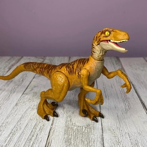 May include: A plastic toy dinosaur, a Velociraptor, in shades of yellow and brown. The dinosaur has a detailed design with visible claws, teeth, and textured skin. The toy is posed on a wooden surface.