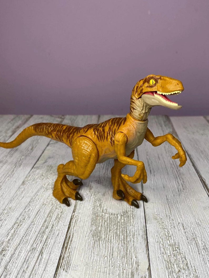 May include: A plastic toy dinosaur, a Velociraptor, in shades of yellow and brown. The dinosaur has a detailed design with visible claws, teeth, and textured skin. The toy is posed on a wooden surface.