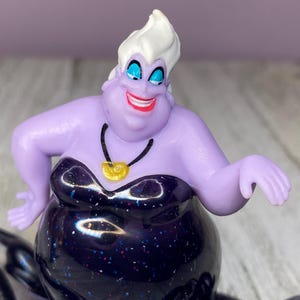 May include: A figurine of Ursula, the sea witch, from Disney's The Little Mermaid. The figure has a purple complexion, white hair, and a black dress with glitter. She wears a gold shell necklace and has a wide, red smile.