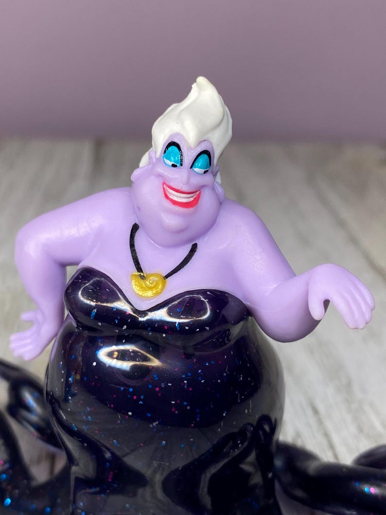 May include: A figurine of Ursula, the sea witch, from Disney's The Little Mermaid. The figure has a purple complexion, white hair, and a black dress with glitter. She wears a gold shell necklace and has a wide, red smile.