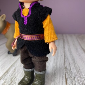 K&ouml;nnte beinhalten: Puppe einer Figur aus dem Animationsfilm Frozen. Die Puppe tr&auml;gt eine schwarze Weste mit lila Besatz &uuml;ber einem gelben Pullover, braune Hosen und gr&uuml;ne Stiefel. Eine Rentierfigur ist teilweise im Hintergrund zu sehen.