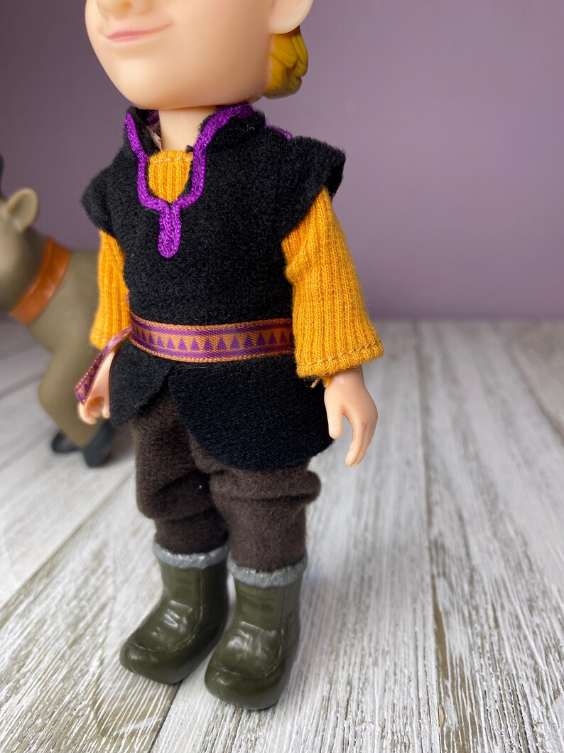 K&ouml;nnte beinhalten: Puppe einer Figur aus dem Animationsfilm Frozen. Die Puppe tr&auml;gt eine schwarze Weste mit lila Besatz &uuml;ber einem gelben Pullover, braune Hosen und gr&uuml;ne Stiefel. Eine Rentierfigur ist teilweise im Hintergrund zu sehen.