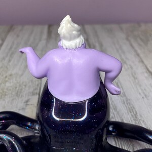 May include: A figurine of Ursula, the sea witch, from Disney's The Little Mermaid. The figure has a purple body, white hair, and a black skirt with glitter. The tentacles are black and extend outward.