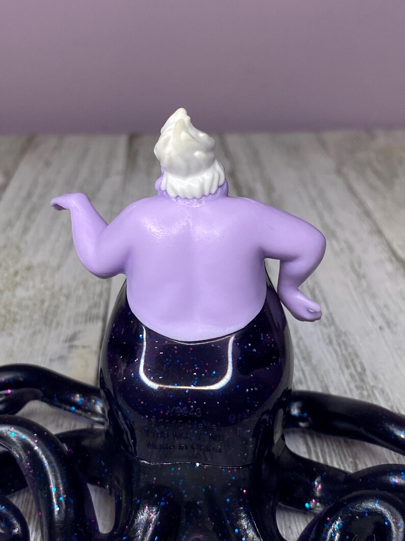 May include: A figurine of Ursula, the sea witch, from Disney's The Little Mermaid. The figure has a purple body, white hair, and a black skirt with glitter. The tentacles are black and extend outward.