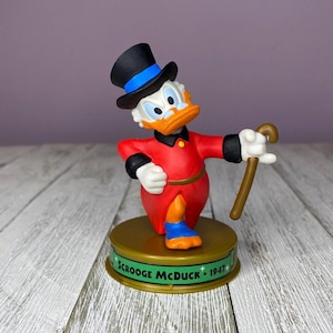 May include: A figurine of Scrooge McDuck, a cartoon character, wearing a red coat, black top hat, and blue shoes. He holds a cane and stands on a base that reads "Scrooge McDuck 1947". The figurine has a white face and an orange beak.
