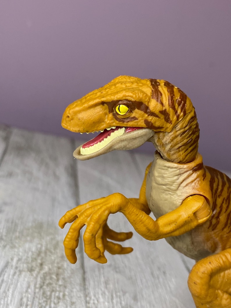 May include: A detailed close-up of a Velociraptor toy. The dinosaur is primarily yellow with brown stripes and has a detailed head, open mouth, and articulated arms. The eyes are yellow, and the mouth is pink.
