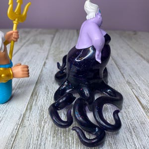 May include: A purple Ursula figurine with a black, glittery octopus body. The figurine has white hair and is posed on a wooden surface. A gold trident is held by a blue and gold figurine in the background.