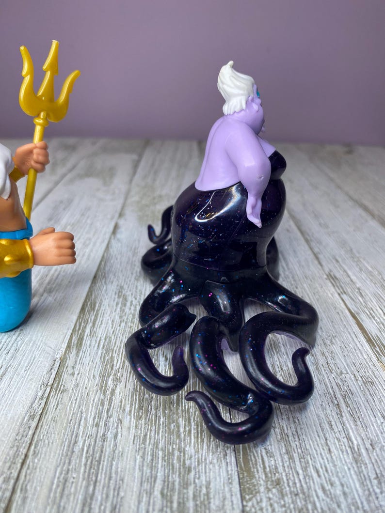 May include: A purple Ursula figurine with a black, glittery octopus body. The figurine has white hair and is posed on a wooden surface. A gold trident is held by a blue and gold figurine in the background.