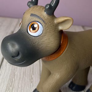 K&ouml;nnte beinhalten: Ein braunes Spielzeugrentier mit gro&szlig;en, ausdrucksstarken Augen und dunkelgrauen Geweihen. Das Rentier hat eine schwarze Nase und einen orangefarbenen Kragen. Das Spielzeug besteht aus Kunststoff und hat eine detaillierte, strukturierte Oberfl&auml;che.