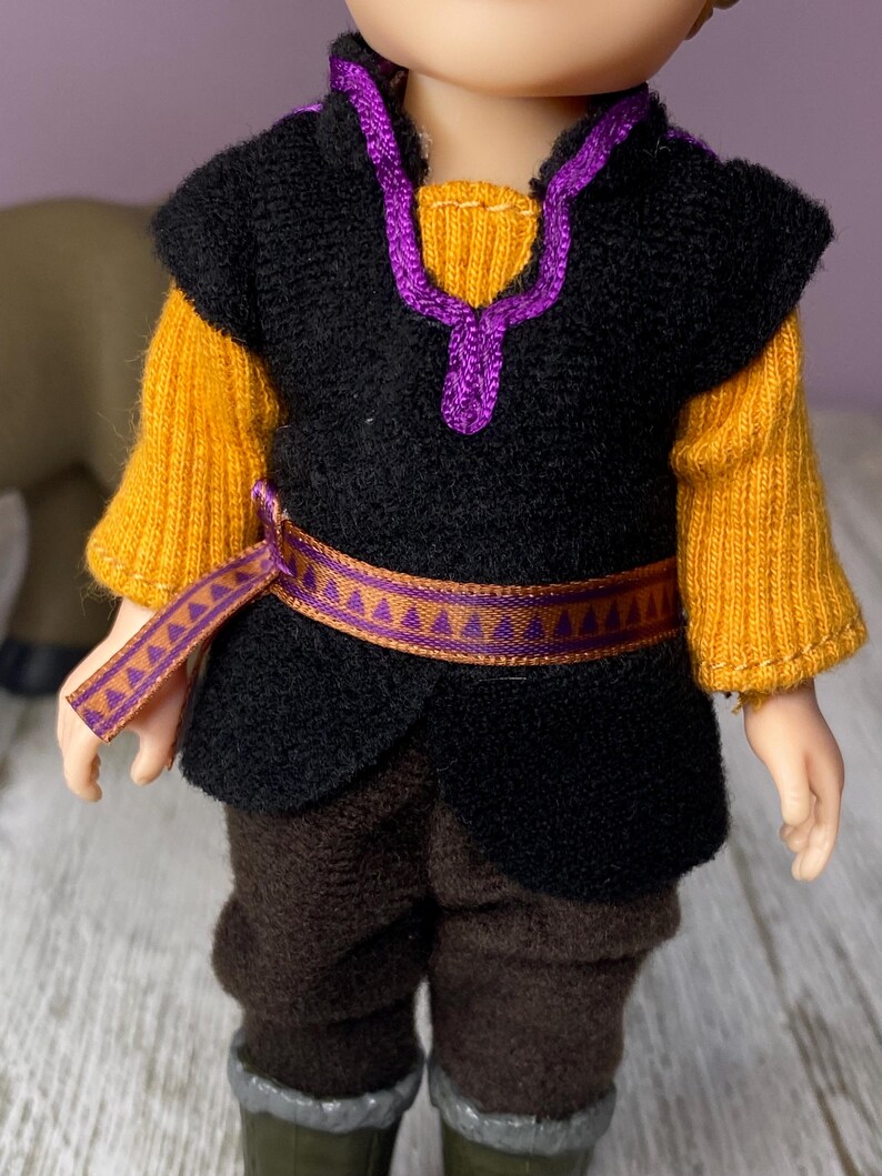 K&ouml;nnte beinhalten: Puppe in einem Kost&uuml;m mit schwarzer Weste, gelben &Auml;rmeln und brauner Hose. Die Weste hat eine lila Verzierung und einen passenden G&uuml;rtel. Das Outfit der Puppe erinnert an eine Figur aus einem beliebten Animationsfilm.