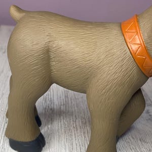 K&ouml;nnte beinhalten: Ein braunes Spielzeugtier mit einem orangefarbenen Halsband. Das Spielzeug hat eine strukturierte Oberfl&auml;che und schwarze Hufe. Das Tier steht auf einer Holzoberfl&auml;che.