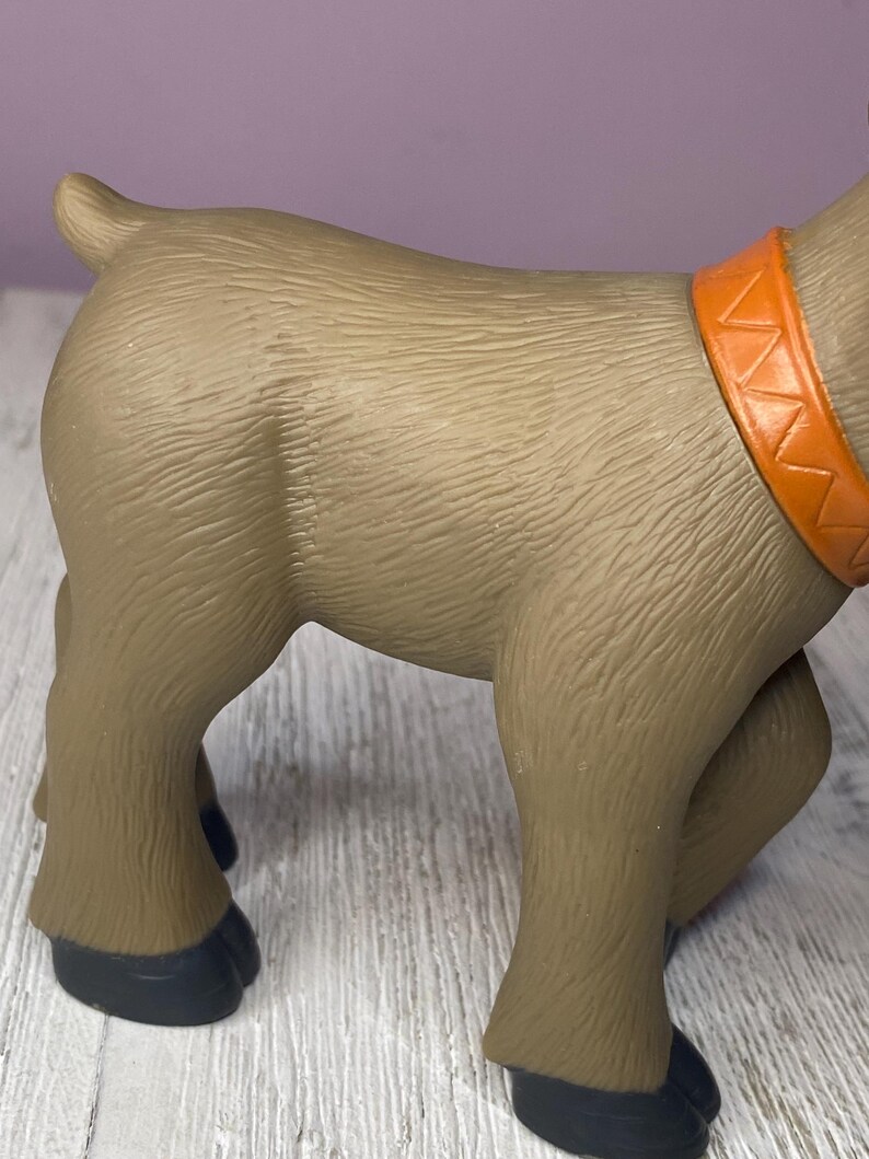 K&ouml;nnte beinhalten: Ein braunes Spielzeugtier mit einem orangefarbenen Halsband. Das Spielzeug hat eine strukturierte Oberfl&auml;che und schwarze Hufe. Das Tier steht auf einer Holzoberfl&auml;che.