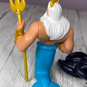 May include: A plastic figurine of a sea king, viewed from the back. The figure has a white beard and hair, a gold crown, and gold armbands. He holds a gold trident and has a blue tail. The figure is on a light-colored surface.