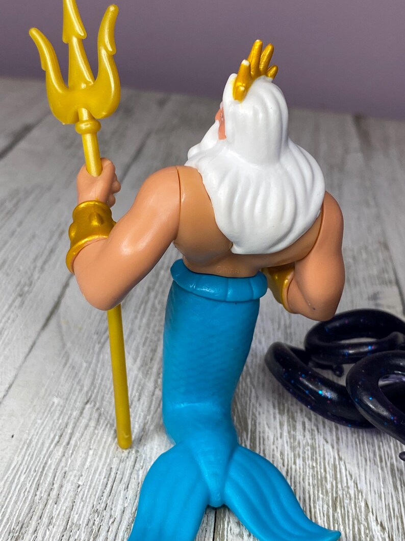 May include: A plastic figurine of a sea king, viewed from the back. The figure has a white beard and hair, a gold crown, and gold armbands. He holds a gold trident and has a blue tail. The figure is on a light-colored surface.