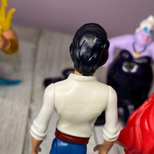 May include: A collection of Disney figurines featuring Prince Eric with black hair, a white shirt, blue pants, and a red belt. The back of the shirt has text that reads "&copy;2013 Mattel". Other figurines include Ursula and Ariel.