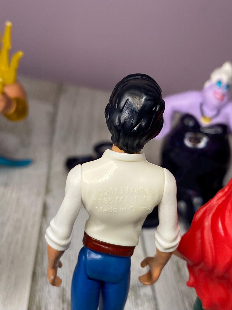 May include: A collection of Disney figurines featuring Prince Eric with black hair, a white shirt, blue pants, and a red belt. The back of the shirt has text that reads "&copy;2013 Mattel". Other figurines include Ursula and Ariel.