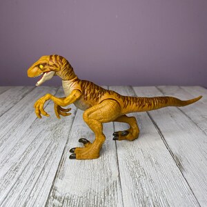 May include: A detailed, yellow and brown dinosaur toy with a long tail, sharp claws, and an open mouth. The toy is posed on a wooden surface against a purple background. The dinosaur has a textured skin pattern.