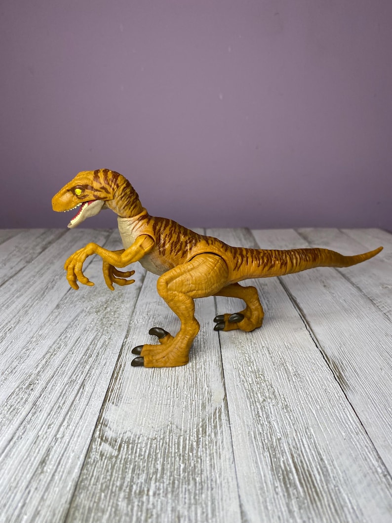 May include: A detailed, yellow and brown dinosaur toy with a long tail, sharp claws, and an open mouth. The toy is posed on a wooden surface against a purple background. The dinosaur has a textured skin pattern.
