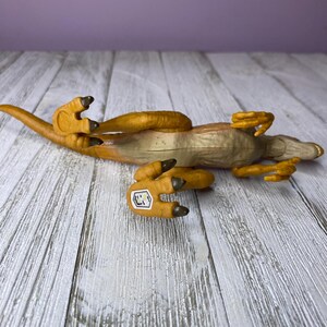 May include: A toy dinosaur, a Velociraptor, lies on its back. The toy is primarily tan and brown, with a lighter underside. The dinosaur's claws and parts of its body are a darker brown. A small sticker is visible on one of its feet.