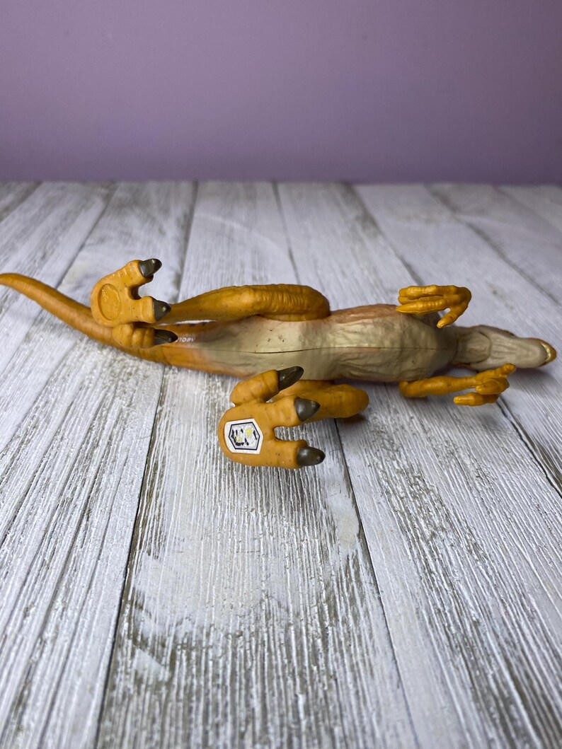 May include: A toy dinosaur, a Velociraptor, lies on its back. The toy is primarily tan and brown, with a lighter underside. The dinosaur's claws and parts of its body are a darker brown. A small sticker is visible on one of its feet.