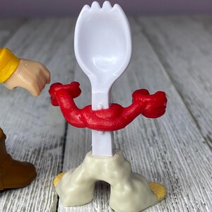May include: A white plastic spork with a red muscular arm design around the handle. The spork is attached to a beige base with yellow accents. A cartoon character's arm and boot are visible.