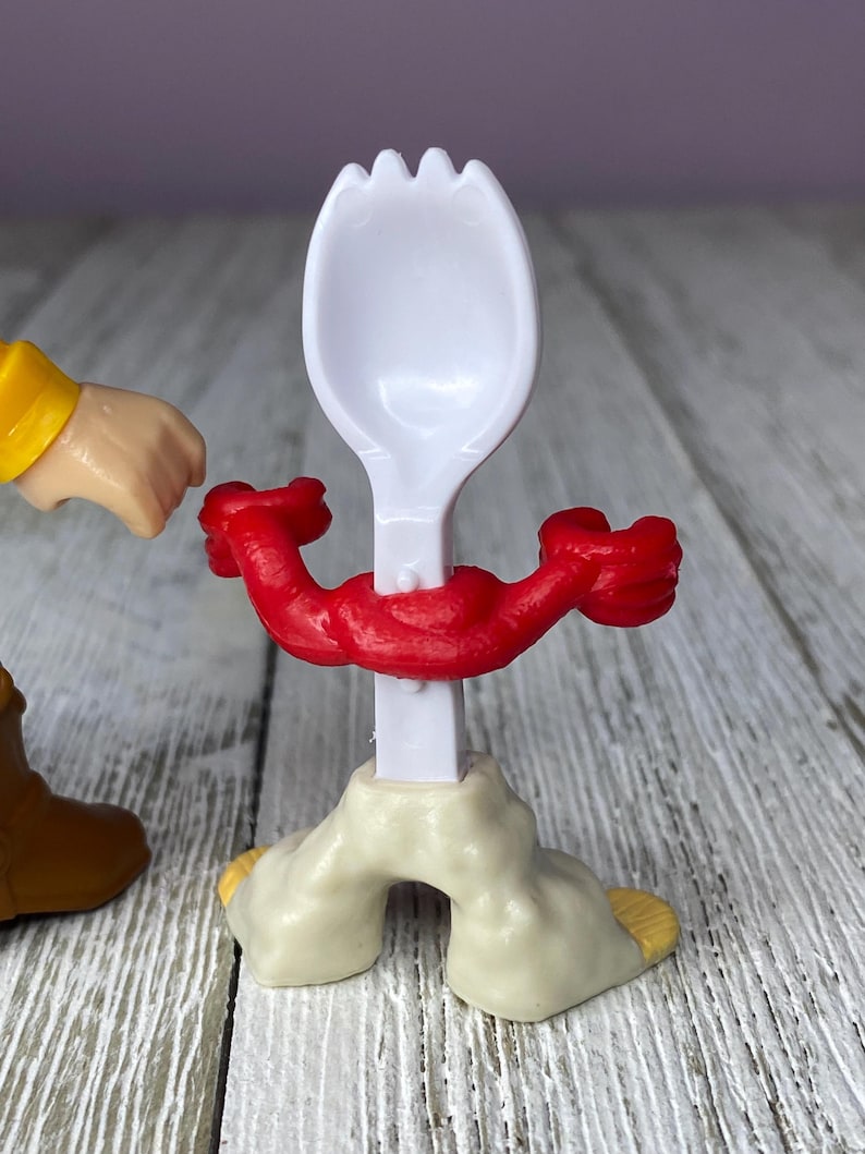 May include: A white plastic spork with a red muscular arm design around the handle. The spork is attached to a beige base with yellow accents. A cartoon character's arm and boot are visible.