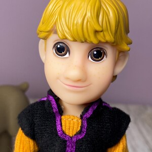 K&ouml;nnte beinhalten: Nahaufnahme einer Puppe mit gelbem Haar und braunen Augen. Die Puppe tr&auml;gt eine schwarze Weste mit lila Besatz &uuml;ber einem gelben Pullover. Die Puppe hat einen l&auml;chelnden Gesichtsausdruck und Sommersprossen.