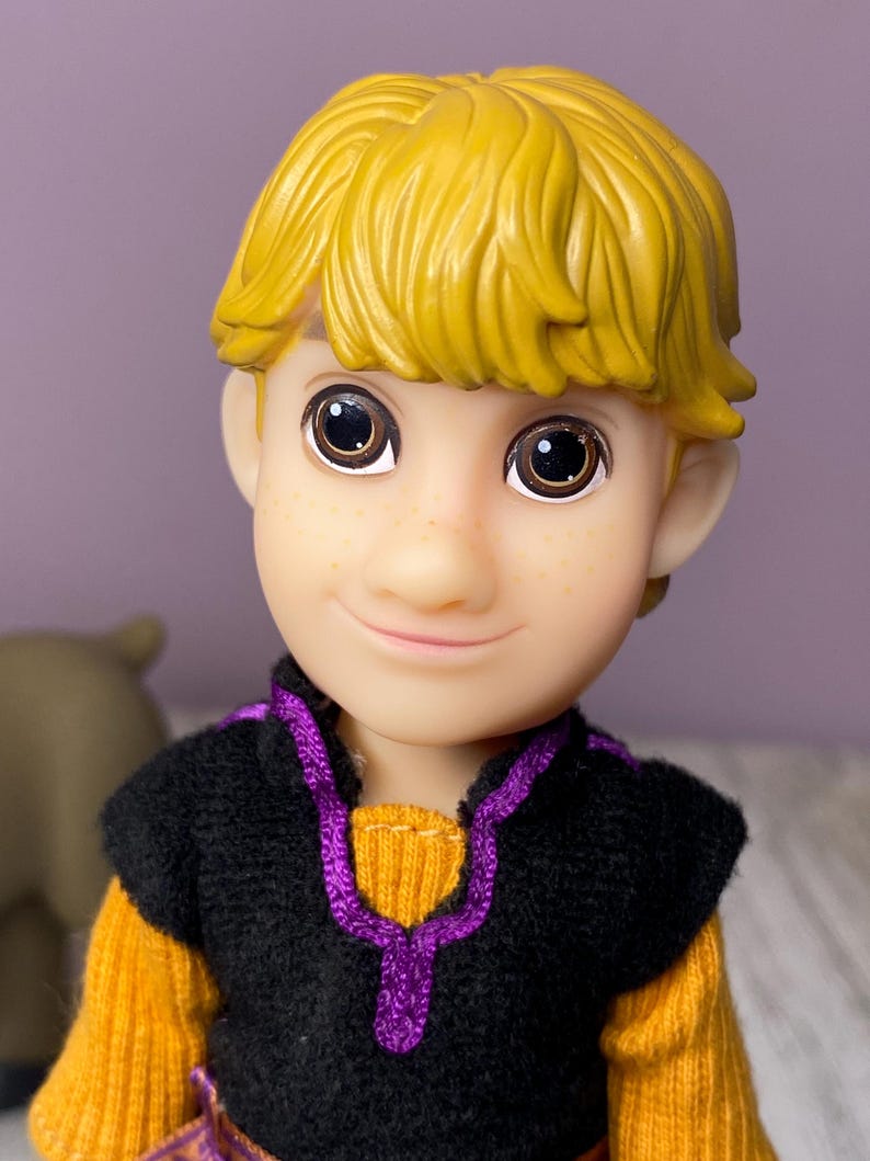 K&ouml;nnte beinhalten: Nahaufnahme einer Puppe mit gelbem Haar und braunen Augen. Die Puppe tr&auml;gt eine schwarze Weste mit lila Besatz &uuml;ber einem gelben Pullover. Die Puppe hat einen l&auml;chelnden Gesichtsausdruck und Sommersprossen.