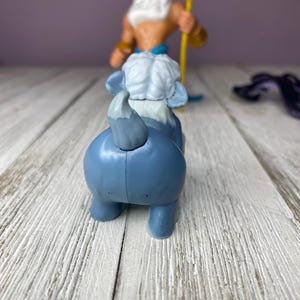 May include: A blue toy elephant with a white head and tail, and a figure of King Triton holding a golden trident in the background. The toys are on a white wooden surface.