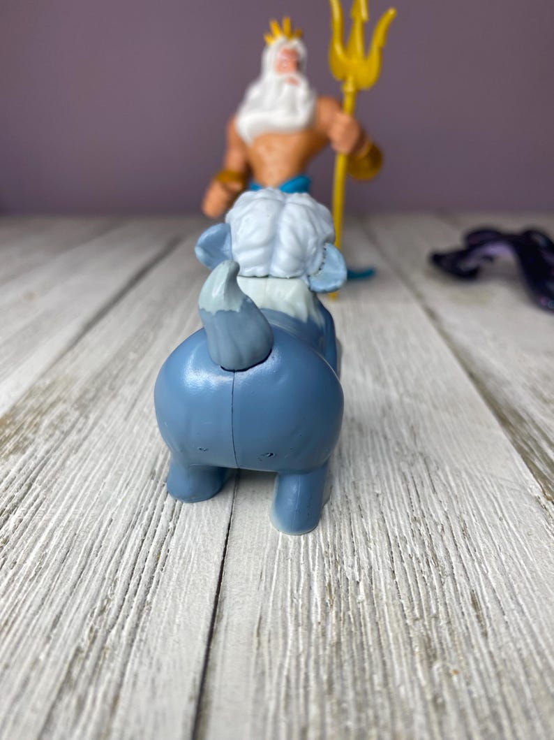 May include: A blue toy elephant with a white head and tail, and a figure of King Triton holding a golden trident in the background. The toys are on a white wooden surface.
