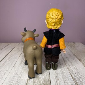 K&ouml;nnte beinhalten: Eine Plastikfigur eines Charakters mit blonden Haaren, der eine schwarze Weste &uuml;ber einem orangefarbenen Pullover, braune Hosen und gr&uuml;ne Stiefel tr&auml;gt, neben einer braunen Rentierfigur mit orangefarbenem Halsband.