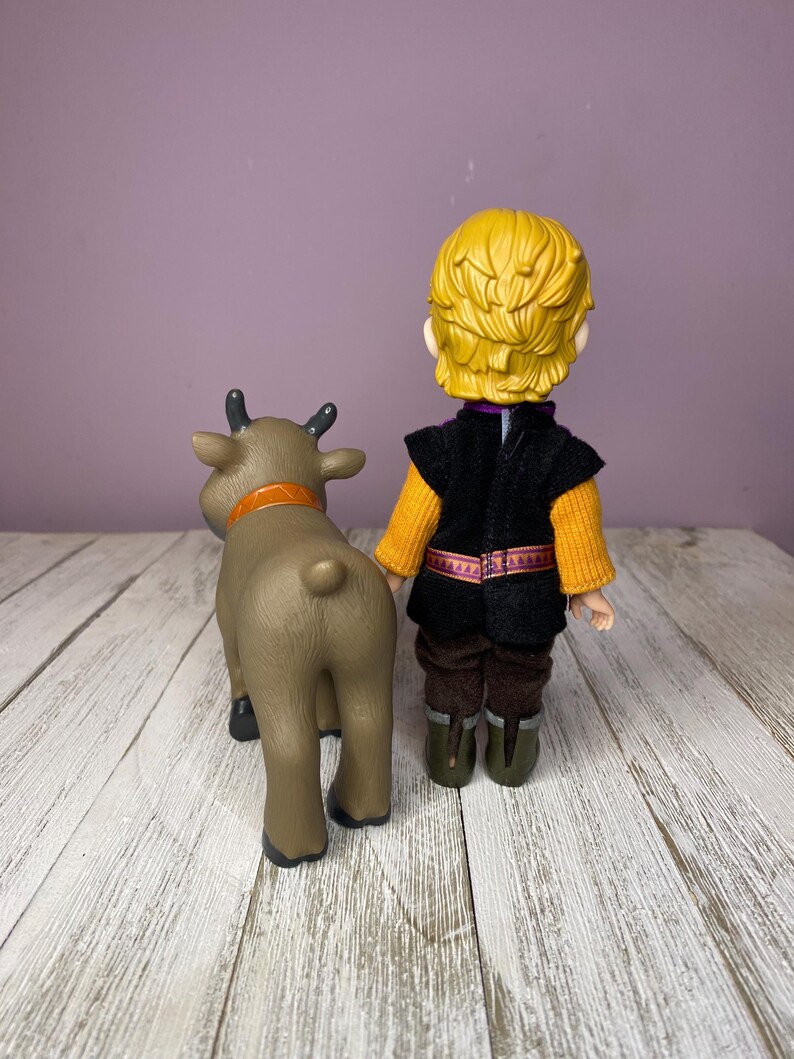 K&ouml;nnte beinhalten: Eine Plastikfigur eines Charakters mit blonden Haaren, der eine schwarze Weste &uuml;ber einem orangefarbenen Pullover, braune Hosen und gr&uuml;ne Stiefel tr&auml;gt, neben einer braunen Rentierfigur mit orangefarbenem Halsband.