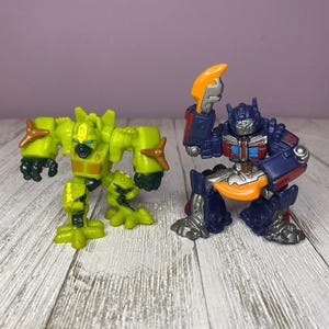 May include: Two toy robot figures. One is lime green with brown accents, the other is blue, red, and silver. Both are posed on a light wood-grain surface against a purple background.