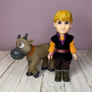 K&ouml;nnte beinhalten: Eine Puppe eines jungen Mannes mit blonden Haaren, der eine schwarze Weste &uuml;ber einem orangefarbenen Hemd, einer braunen Hose und gr&uuml;nen Stiefeln tr&auml;gt. Eine braune Rentierfigur mit braunem Halsband steht neben der Puppe.