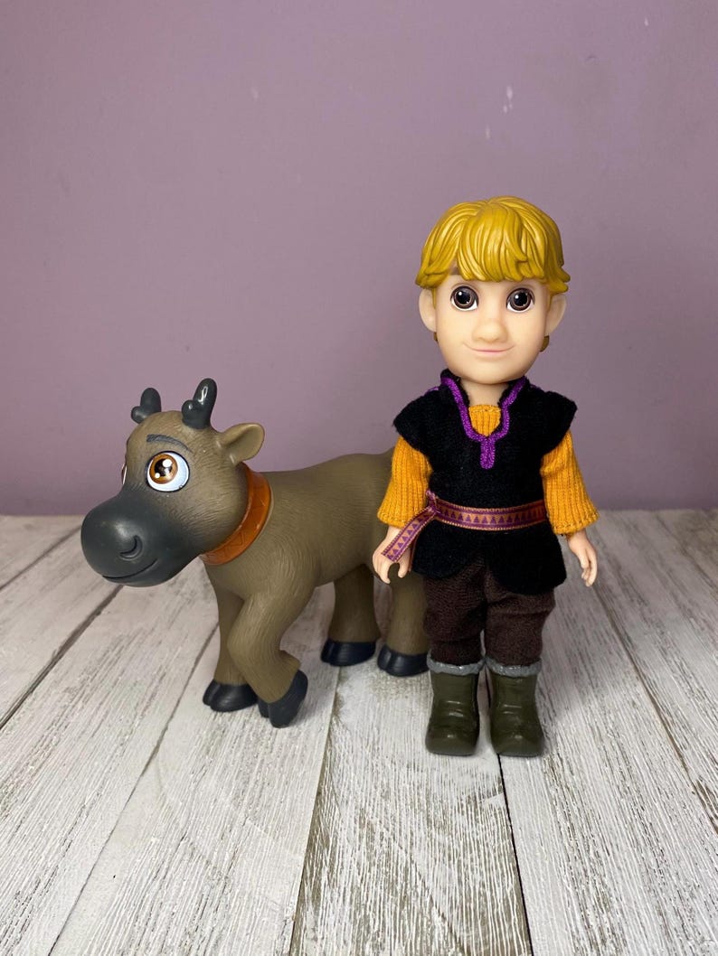 K&ouml;nnte beinhalten: Eine Puppe eines jungen Mannes mit blonden Haaren, der eine schwarze Weste &uuml;ber einem orangefarbenen Hemd, einer braunen Hose und gr&uuml;nen Stiefeln tr&auml;gt. Eine braune Rentierfigur mit braunem Halsband steht neben der Puppe.