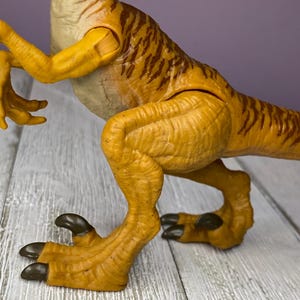 May include: A detailed, close-up shot of a yellow and brown dinosaur toy. The toy features articulated limbs and a textured skin pattern. The dinosaur's claws are dark brown. The toy is positioned on a light gray wooden surface.
