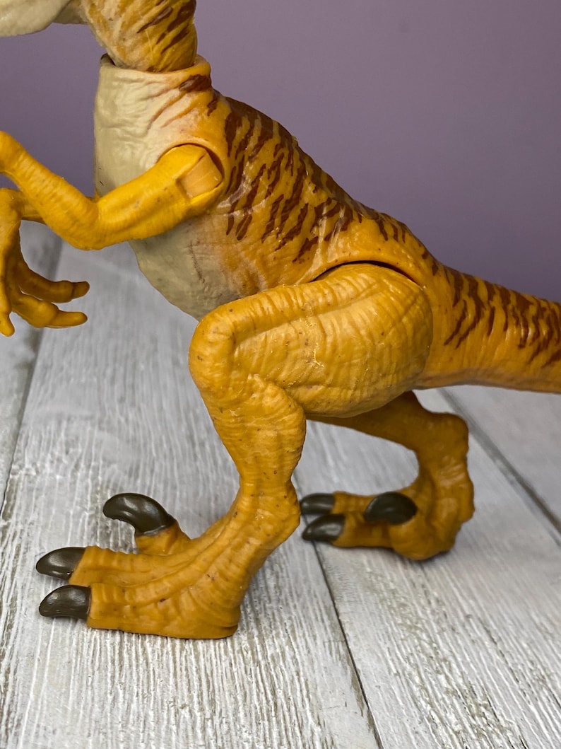 May include: A detailed, close-up shot of a yellow and brown dinosaur toy. The toy features articulated limbs and a textured skin pattern. The dinosaur's claws are dark brown. The toy is positioned on a light gray wooden surface.