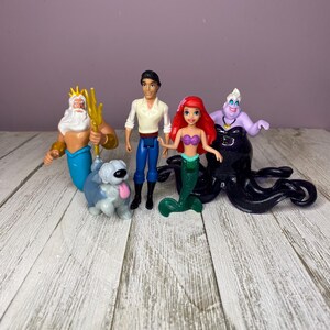 May include: A collection of Disney's The Little Mermaid figurines. The set includes Ariel, Prince Eric, King Triton, Ursula, and a dog. The figurines are brightly colored and made of plastic. The background is a light purple wall and a white wooden surface.
