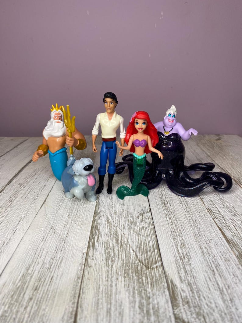 May include: A collection of Disney's The Little Mermaid figurines. The set includes Ariel, Prince Eric, King Triton, Ursula, and a dog. The figurines are brightly colored and made of plastic. The background is a light purple wall and a white wooden surface.