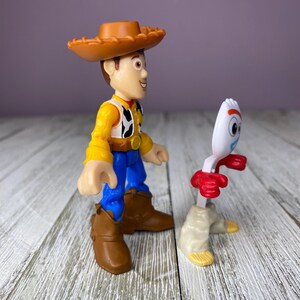 May include: Two Toy Story character figurines. Woody, in a brown cowboy hat, yellow shirt, cow-print vest, blue pants, and brown boots, stands next to Forky, a white spork with red arms and beige legs.