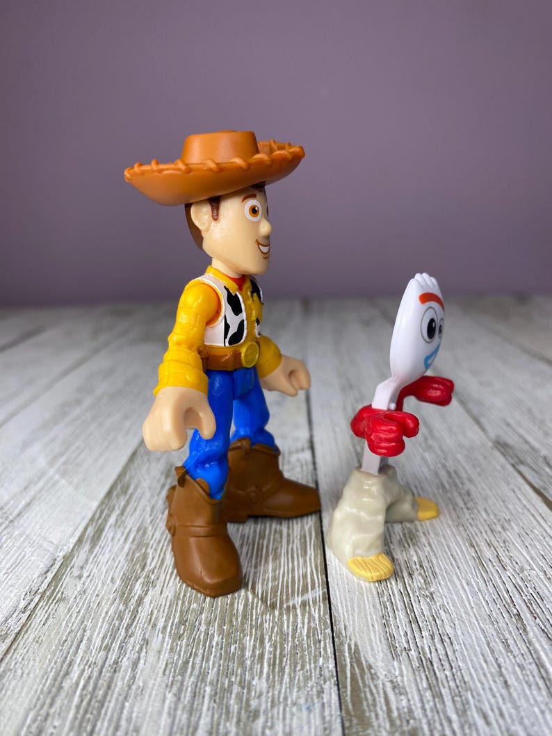 May include: Two Toy Story character figurines. Woody, in a brown cowboy hat, yellow shirt, cow-print vest, blue pants, and brown boots, stands next to Forky, a white spork with red arms and beige legs.