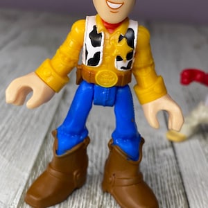 May include: A Woody action figure from the Toy Story franchise. The figure is wearing a yellow shirt, a black and white vest, blue pants, and brown boots. The figure has a sheriff's badge and a brown belt with a gold buckle.